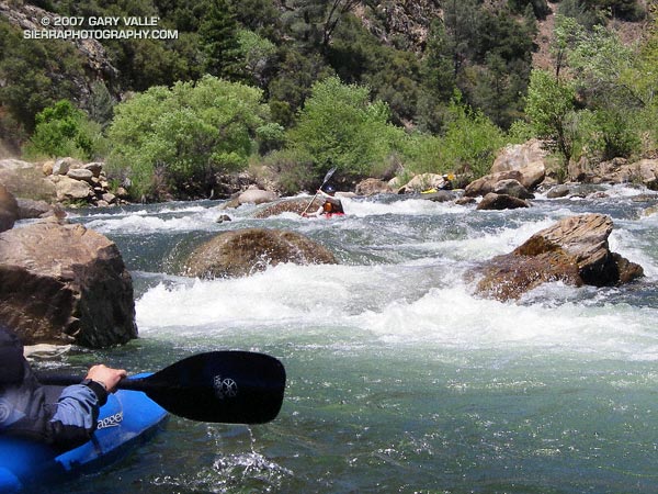 A kayaker waits in an eddy, while two other paddlers work their way through the last third of Limestone rapid, one of the classic class IV rapids on the upper Kern River.